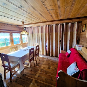 A wooden cabin with a dining area featuring a table and chairs, a red sofa, and a window with curtains. - Mayen Motau - Retour à l'essentiel | La Forclaz (La Forclaz)