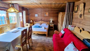 A wooden cabin interior with a bed, a red sofa, a dining table, and a chandelier. - Mayen Motau - Retour à l'essentiel | La Forclaz (La Forclaz)