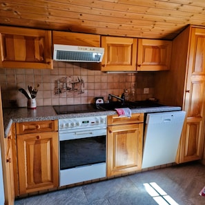 A kitchen with wooden cabinets, a stove, a dishwasher, and a tiled backsplash. - Mayen Motau - Retour à l'essentiel | La Forclaz (La Forclaz)