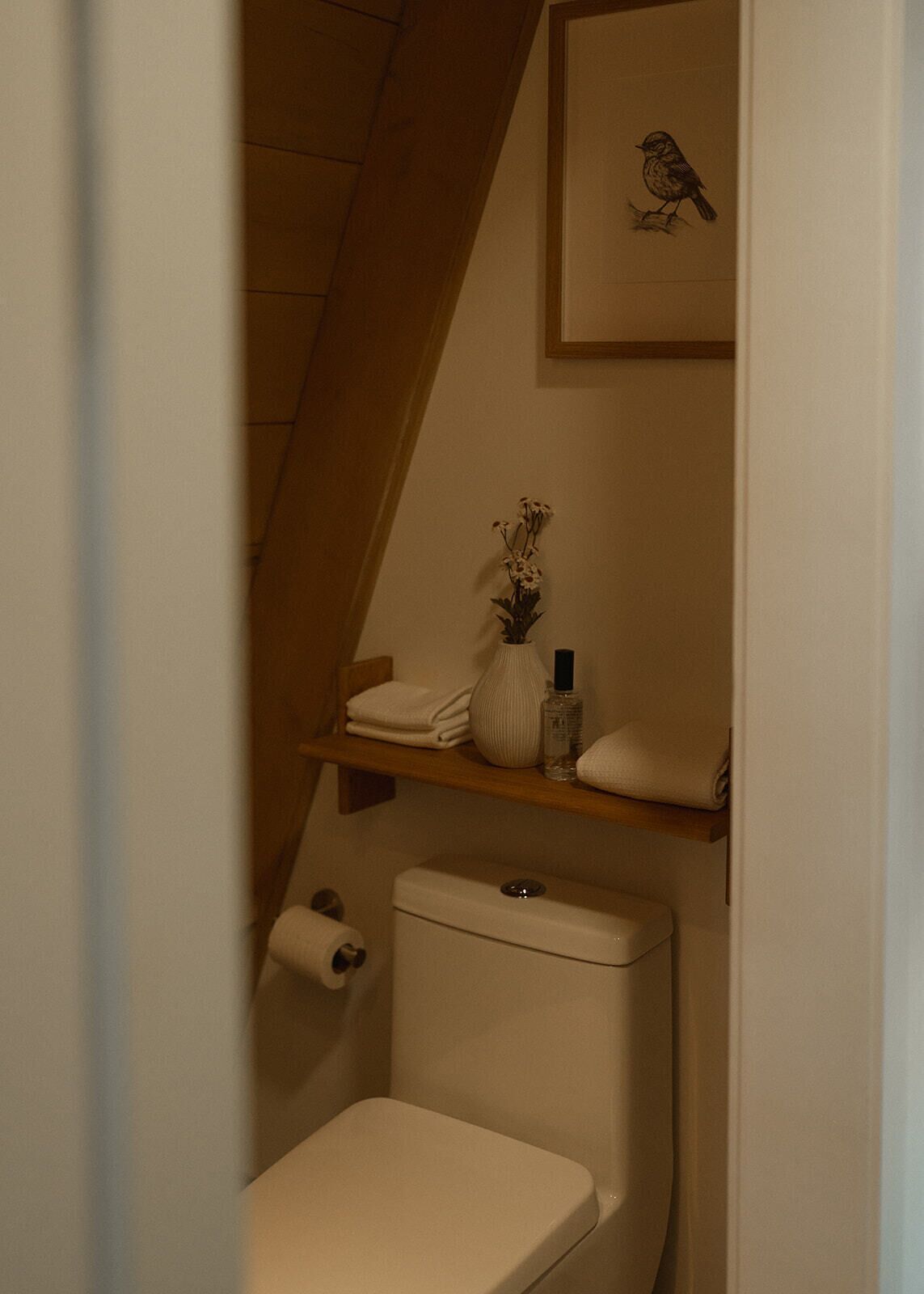 Un baño con inodoro, un rollo de papel higiénico, un estante pequeño con un florero y artículos de aseo, y un cuadro con la imagen de un pájaro en la pared.