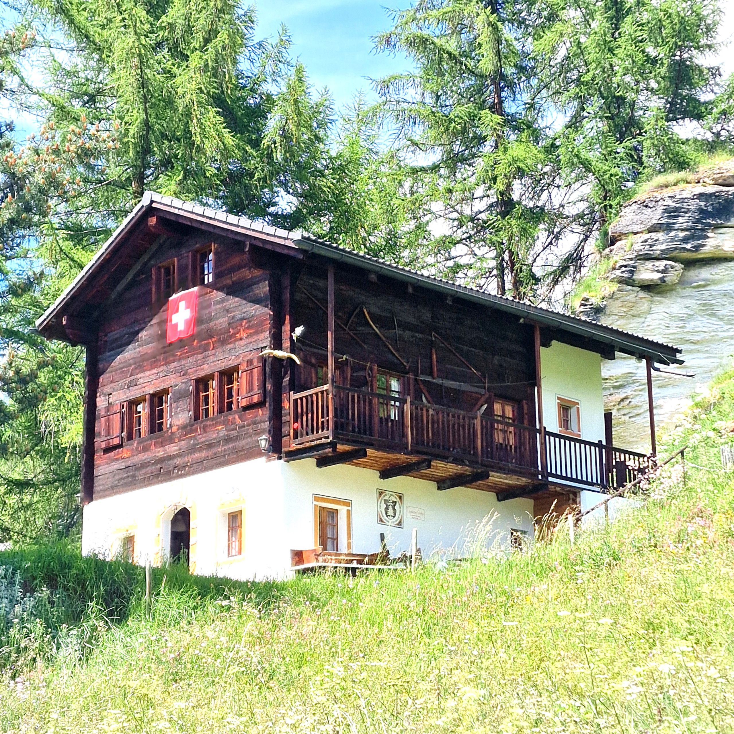 A wooden chalet with a balcony, set against a backdrop of trees and a rocky cliff.
