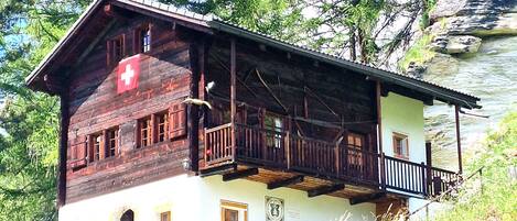 A wooden chalet with a balcony, set against a backdrop of trees and a rocky cliff.