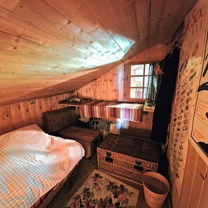 A cozy attic bedroom with wooden walls and a sloped ceiling. There is a bed with a checkered bedspread, a small seating area with a cushioned bench, and a wicker basket.
