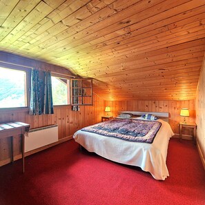 A wooden cabin bedroom with a bed, a window with curtains, a heater, and a lamp on a bedside table.