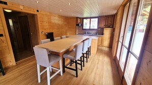 A wooden interior with a long dining table, chairs, and a kitchen area.