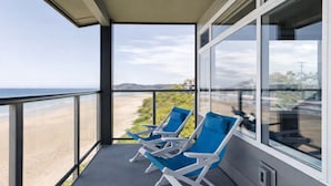 Un balcon avec deux fauteuils bleus, une balustrade en verre et une vue sur la plage.