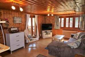 Interior de una cabaña de madera con sofá, televisor, cómoda blanca y mesa de comedor.