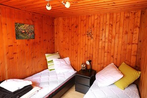 A small, cozy room with wooden walls, two single beds with pillows, a nightstand with a lamp, and a wall hanging.
