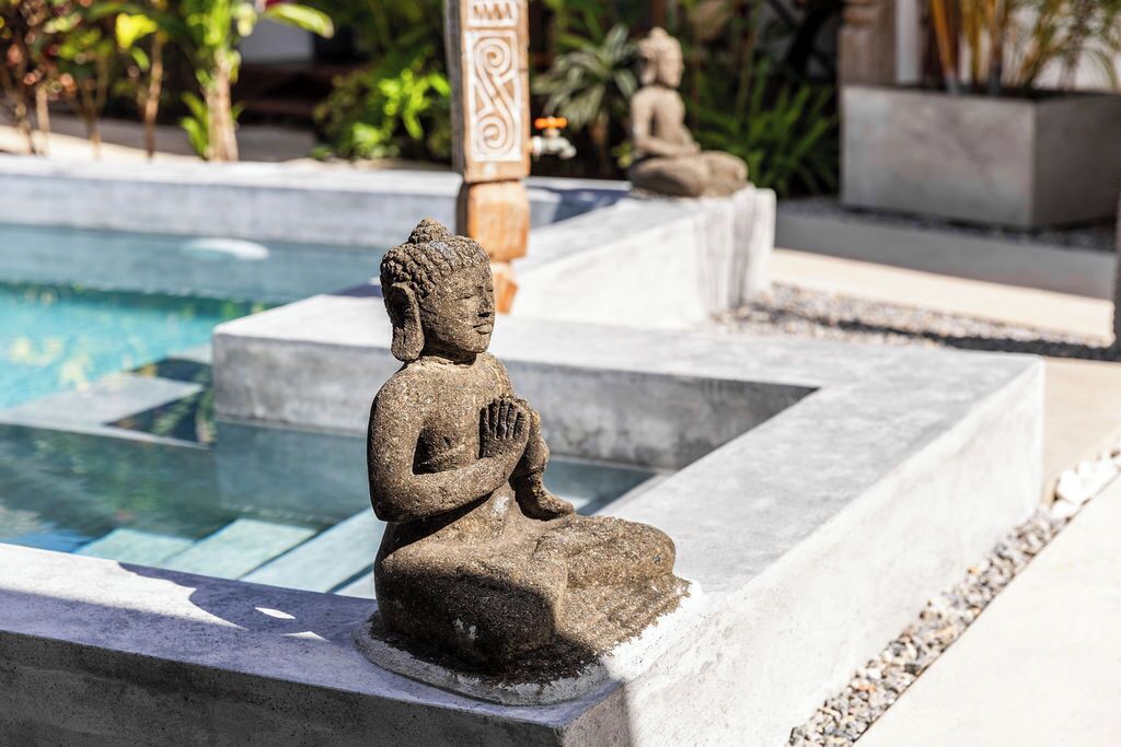 A stone Buddha statue by a poolside, surrounded by a stone pathway and greenery.