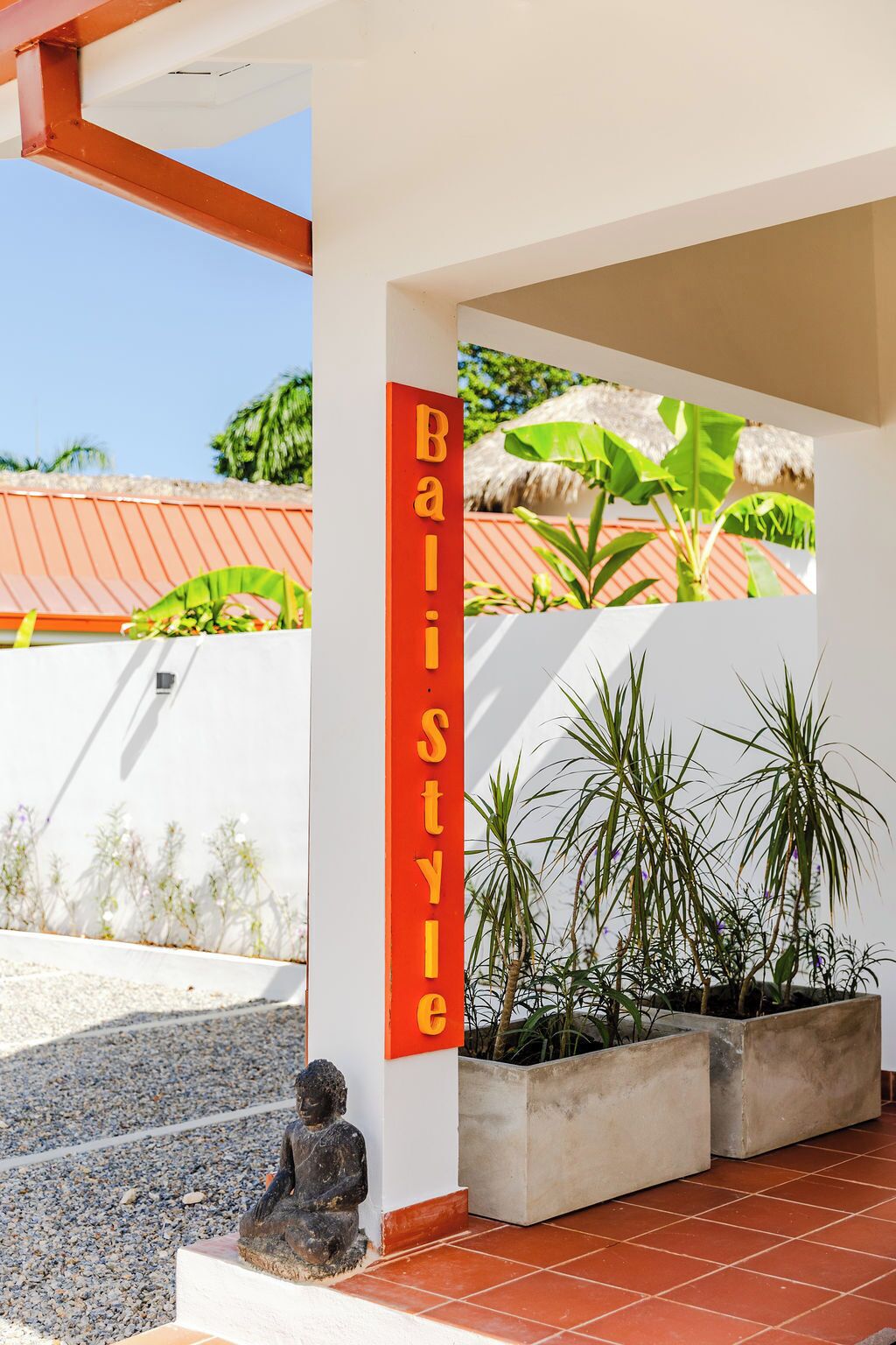 Un bâtiment moderne aux couleurs rouge et blanche, doté d’une grande fenêtre offrant une vue sur un paysage tropical, et d’une petite statue de Bouddha placée à l’avant.