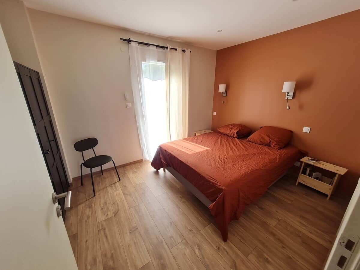 Ein Schlafzimmer mit einem Bett, einem Sessel, einem Nachttisch und einem Fenster mit Vorhängen.