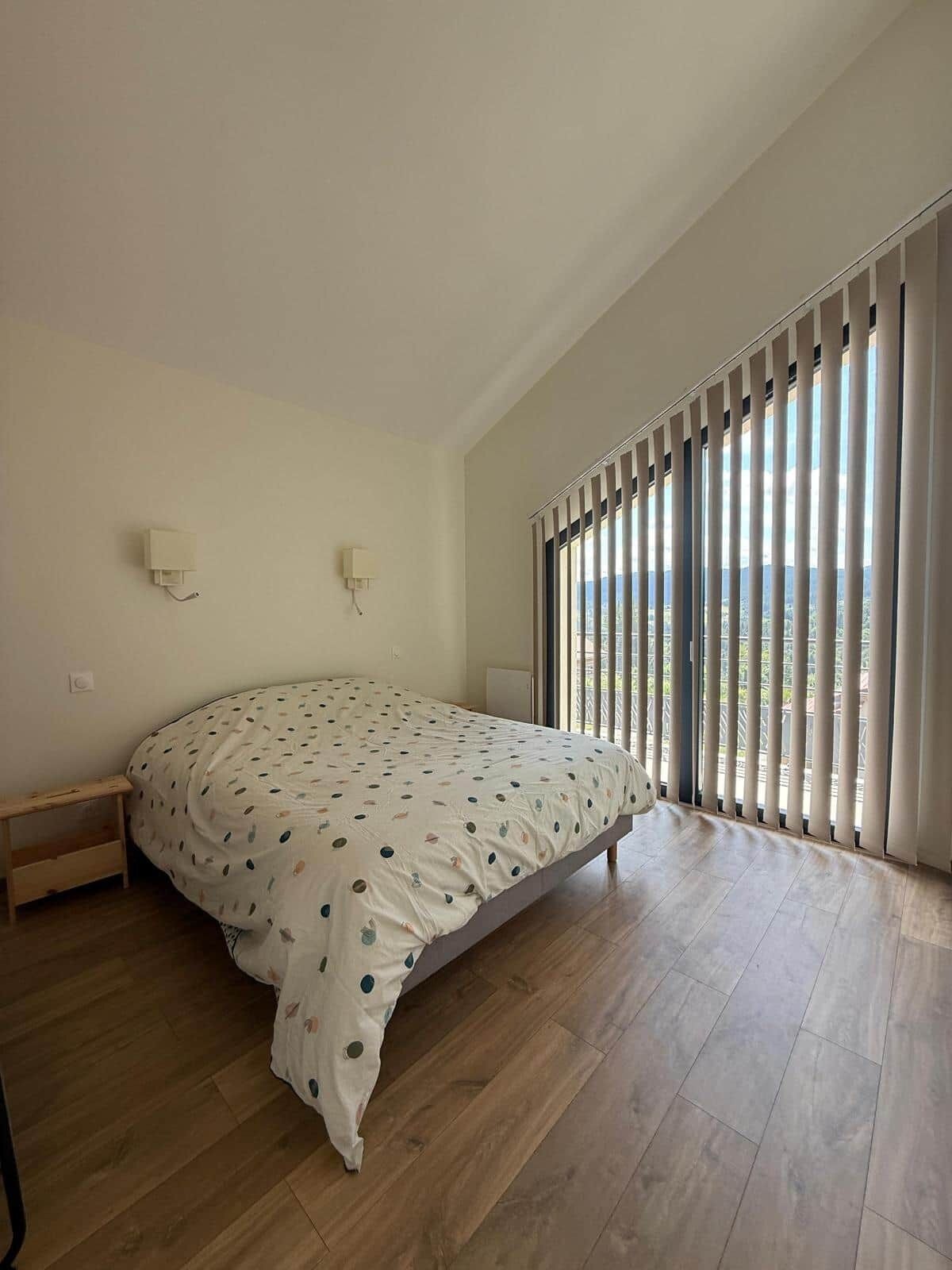 A bedroom with a bed, a nightstand, a wall-mounted lamp, and a large window with blinds.