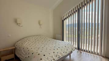 A bedroom with a bed, a nightstand, a wall-mounted lamp, and a large window with blinds.