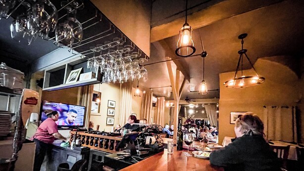 Zona de bar con una barra de madera larga, copas de vino colgando y una pantalla de televisión mostrando un evento deportivo.
