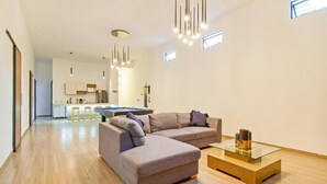 A modern living room with a sectional sofa, a glass coffee table, and pendant lighting. A kitchen with bar stools is visible in the background.