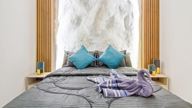 A bed with a grey quilt, two teal pillows, and a purple towel shaped like a swan.