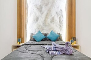 Una cama con un edredón gris, dos almohadas color turquesa y una toalla morada con forma de cisne.