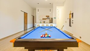 A pool table with balls and cues, a kitchen area in the background, and a hallway leading to a door.