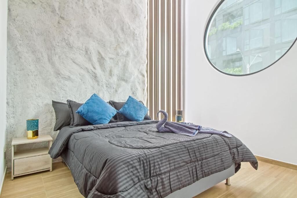 Un dormitorio con cama, mesita de noche, ventana y vistas a edificios.