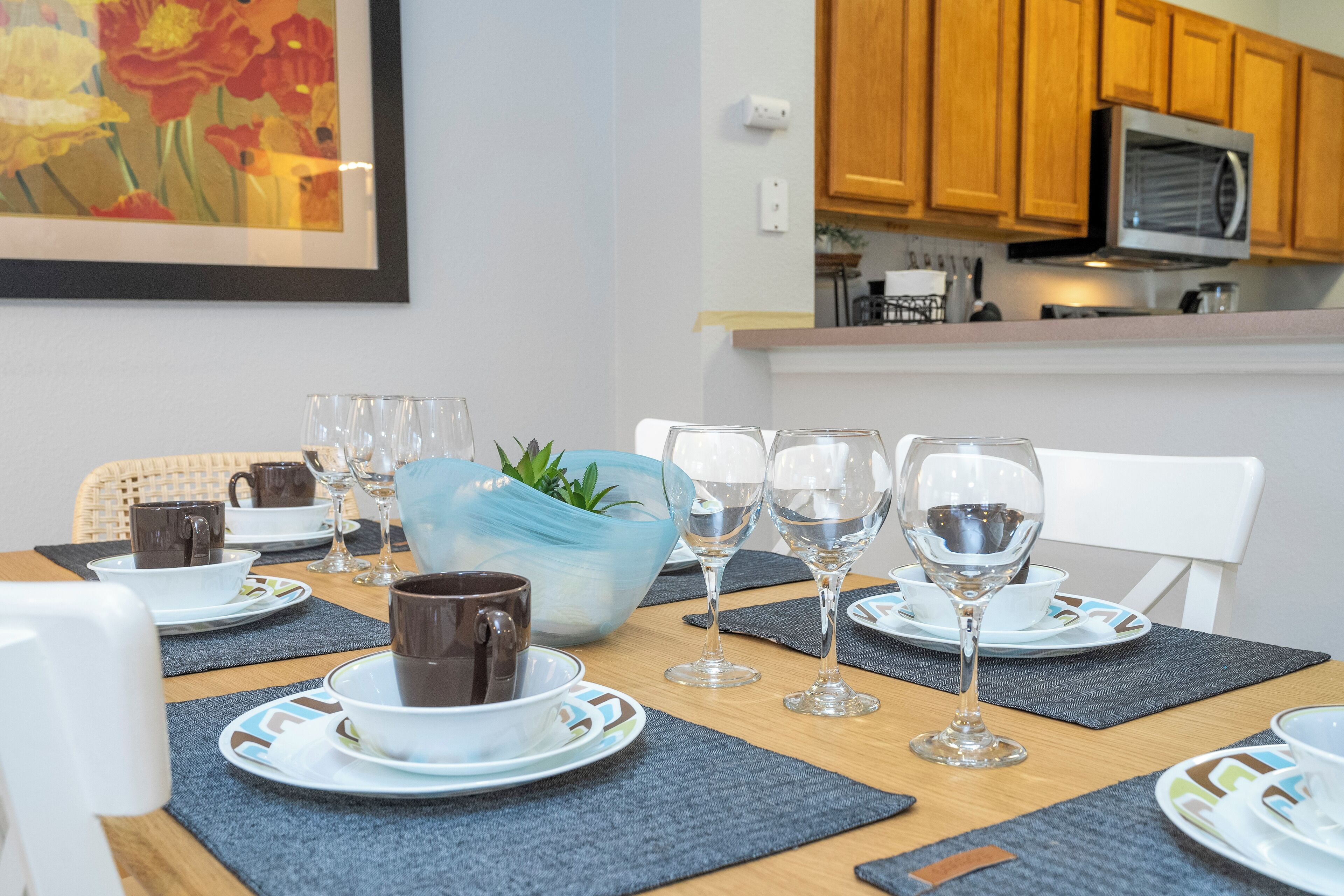Una mesa de comedor dispuesta con platos, vasos y una taza de café, con un microondas y armarios de madera al fondo.