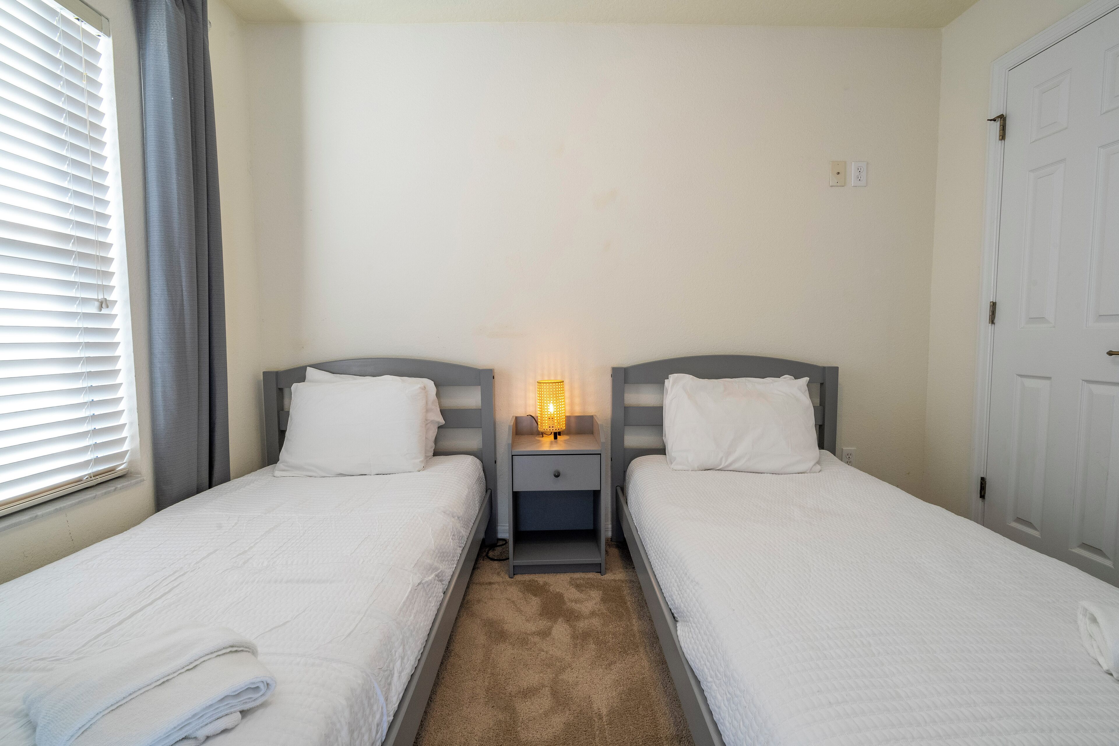 Habitación doble con dos camas, una mesita de noche con lámpara y una ventana con persianas.