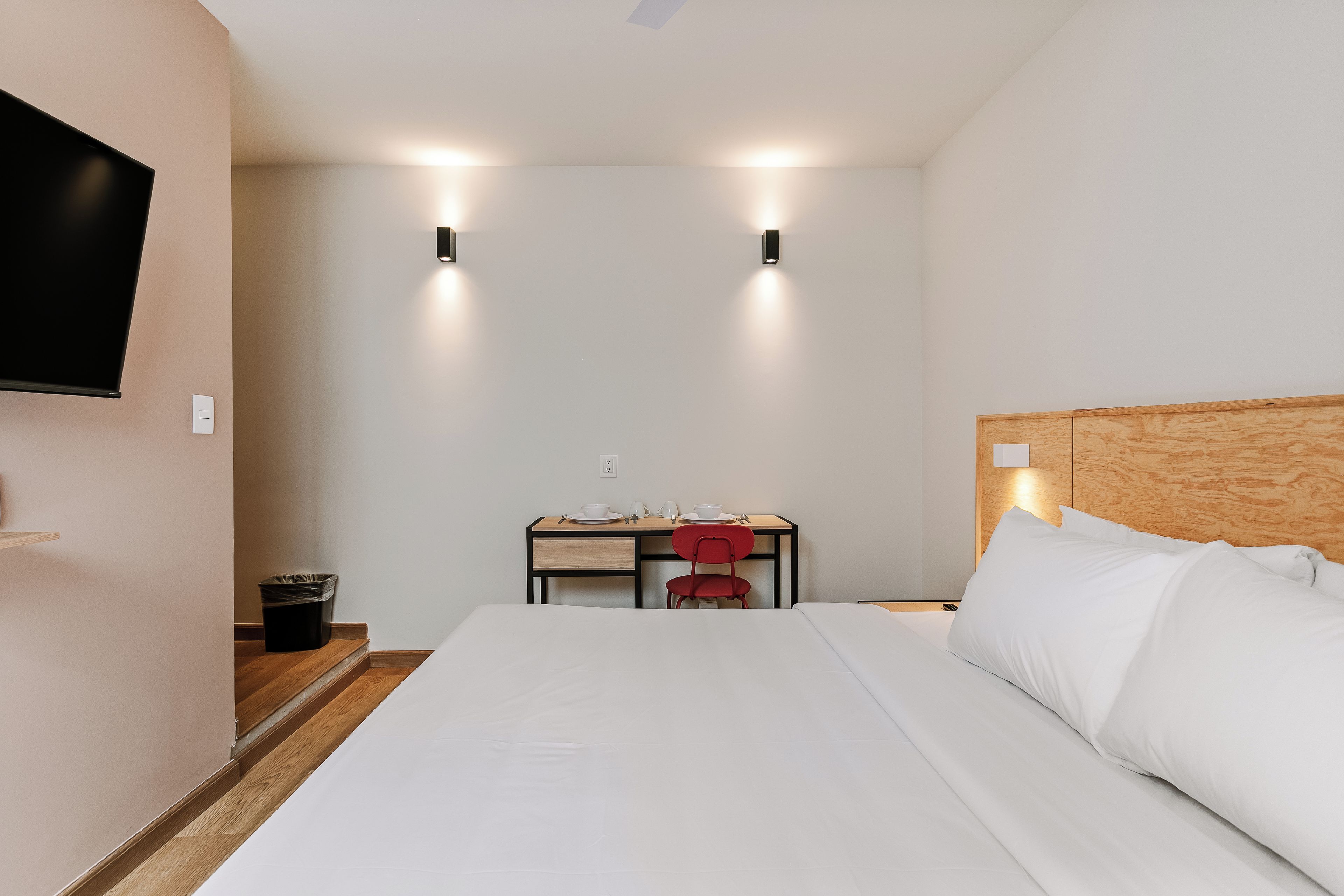 A hotel room with a bed, a desk with a chair, a television mounted on the wall, and a trash bin.