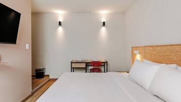 A hotel room with a bed, a desk with a chair, a television mounted on the wall, and a trash bin.