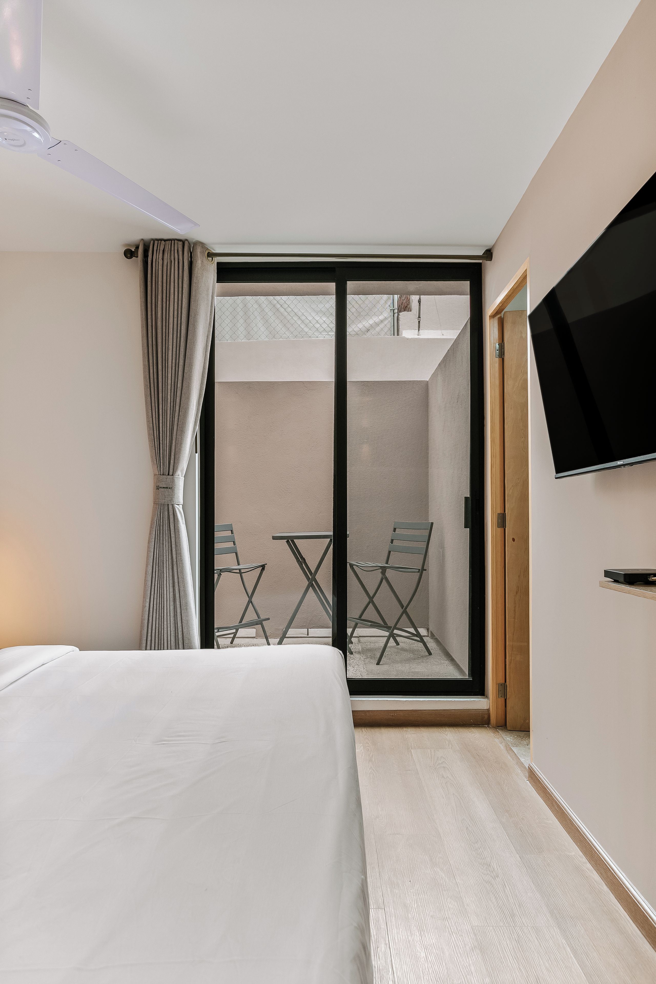 Habitación de hotel con cama, televisor montado en la pared y puerta corrediza de vidrio que da a un área exterior con dos sillas.
