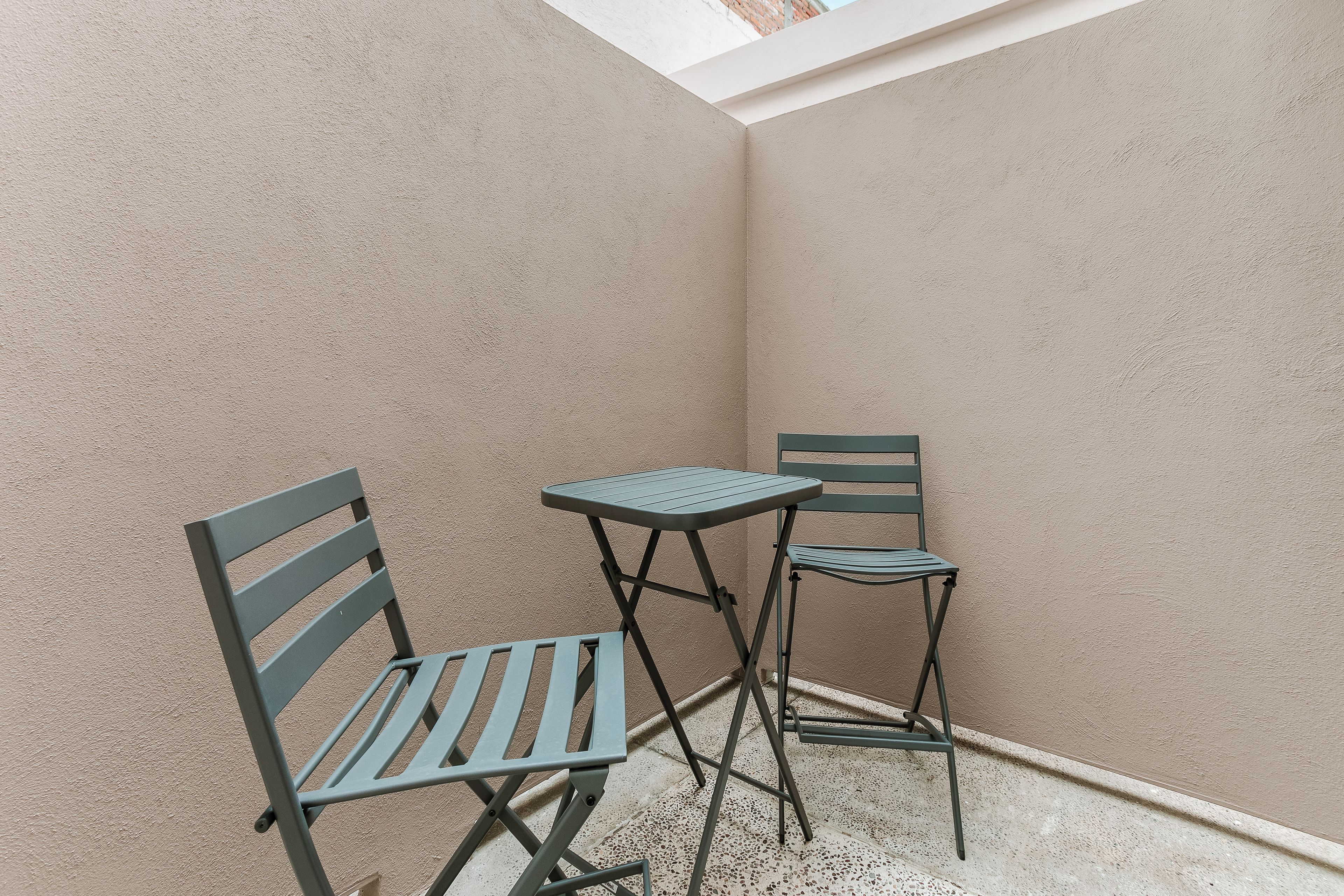 Ein kleiner Sitzbereich im Freien mit zwei Metallstühlen und einem Tisch, vor einer Wand mit strukturierter Oberfläche und geneigter Decke.