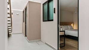 Un pasillo estrecho con una escalera a la izquierda, una puerta con ventanal a la derecha y una cama con cabecera en la habitación contigua.
