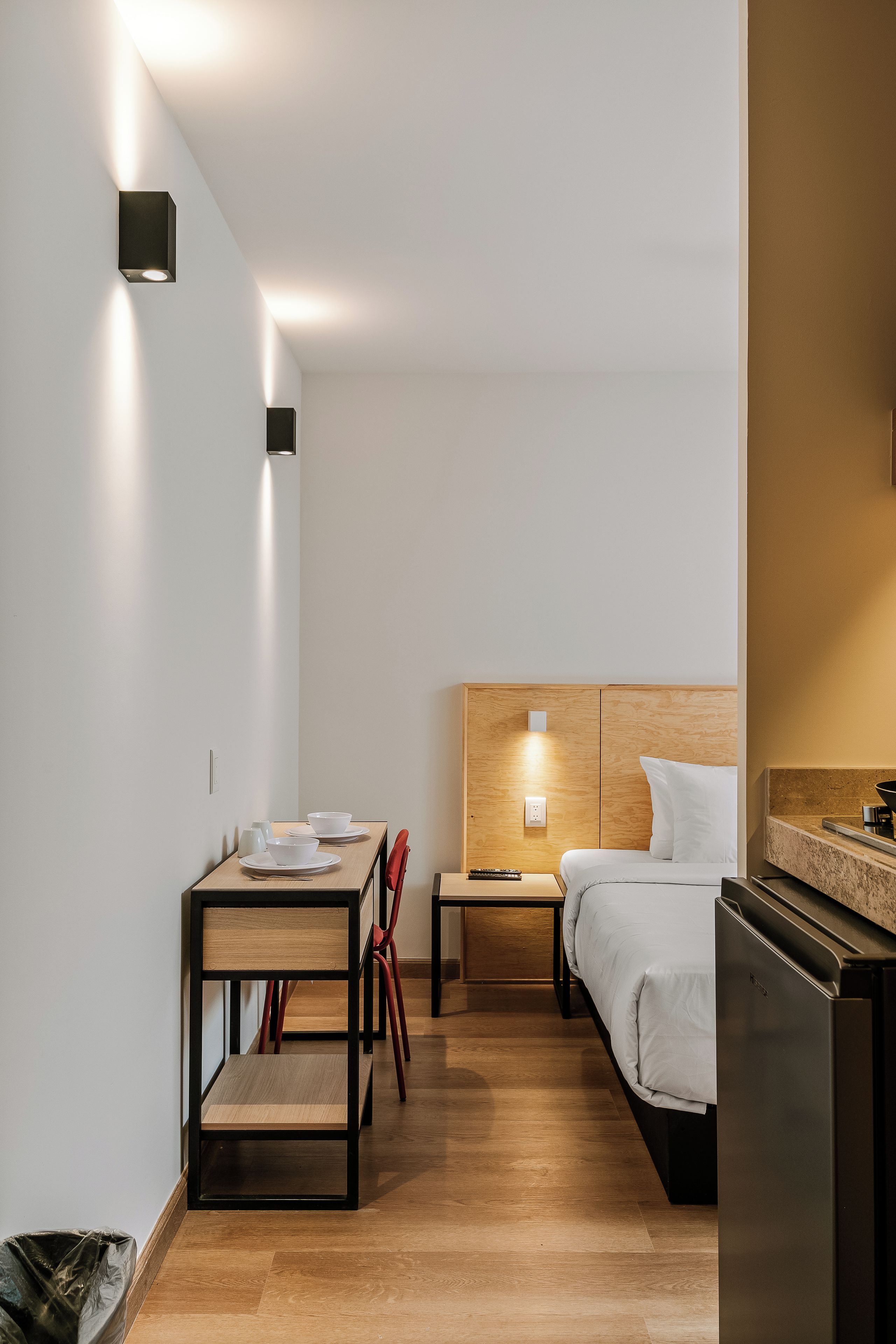 A compact hotel room with a wooden floor, a bed with a headboard, a dining table with chairs, a small table with a remote control, and a kitchenette area.