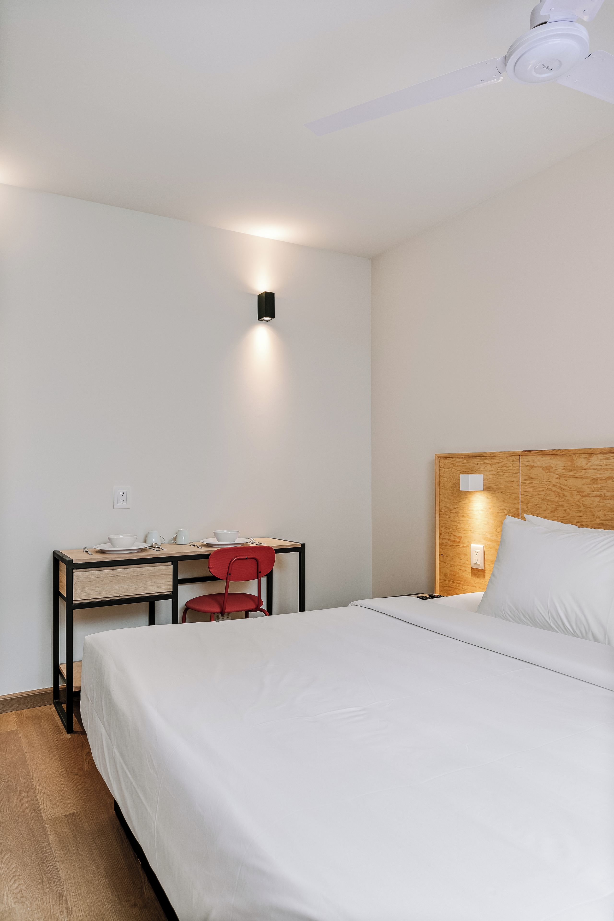 A hotel room with a bed, a desk with a chair, a ceiling fan, and a wall-mounted light.