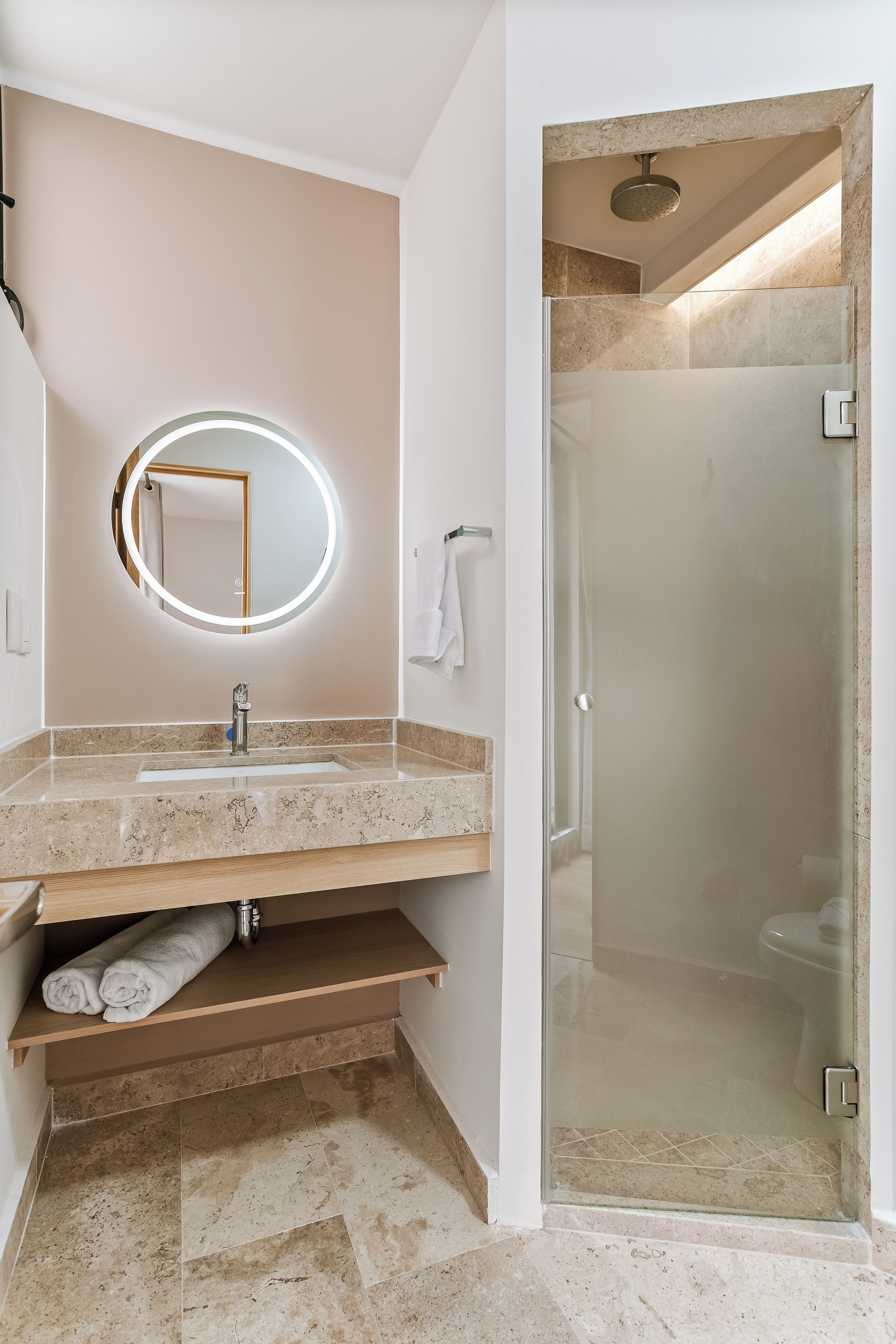 A bathroom with a marble sink, a round mirror with lighting, a towel rack, and a glass-enclosed shower.