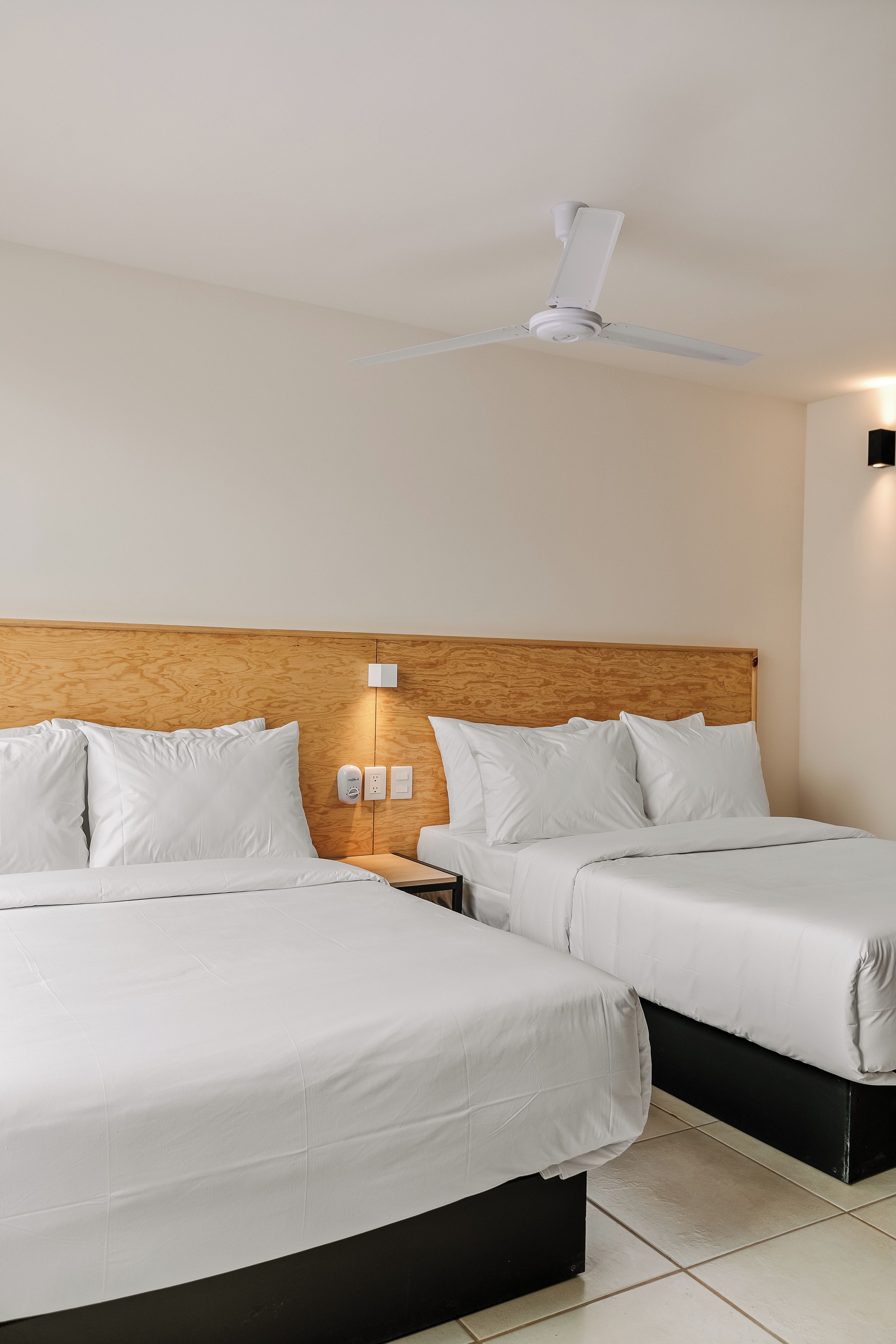 A hotel room with two beds, a wooden headboard, a ceiling fan, and a wall-mounted light fixture.