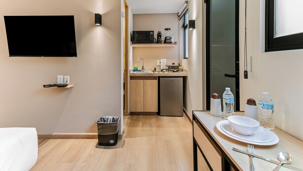 Una habitación de hotel compacta con cama, kitchenette pequeña, televisor y comedor con mesa para dos.