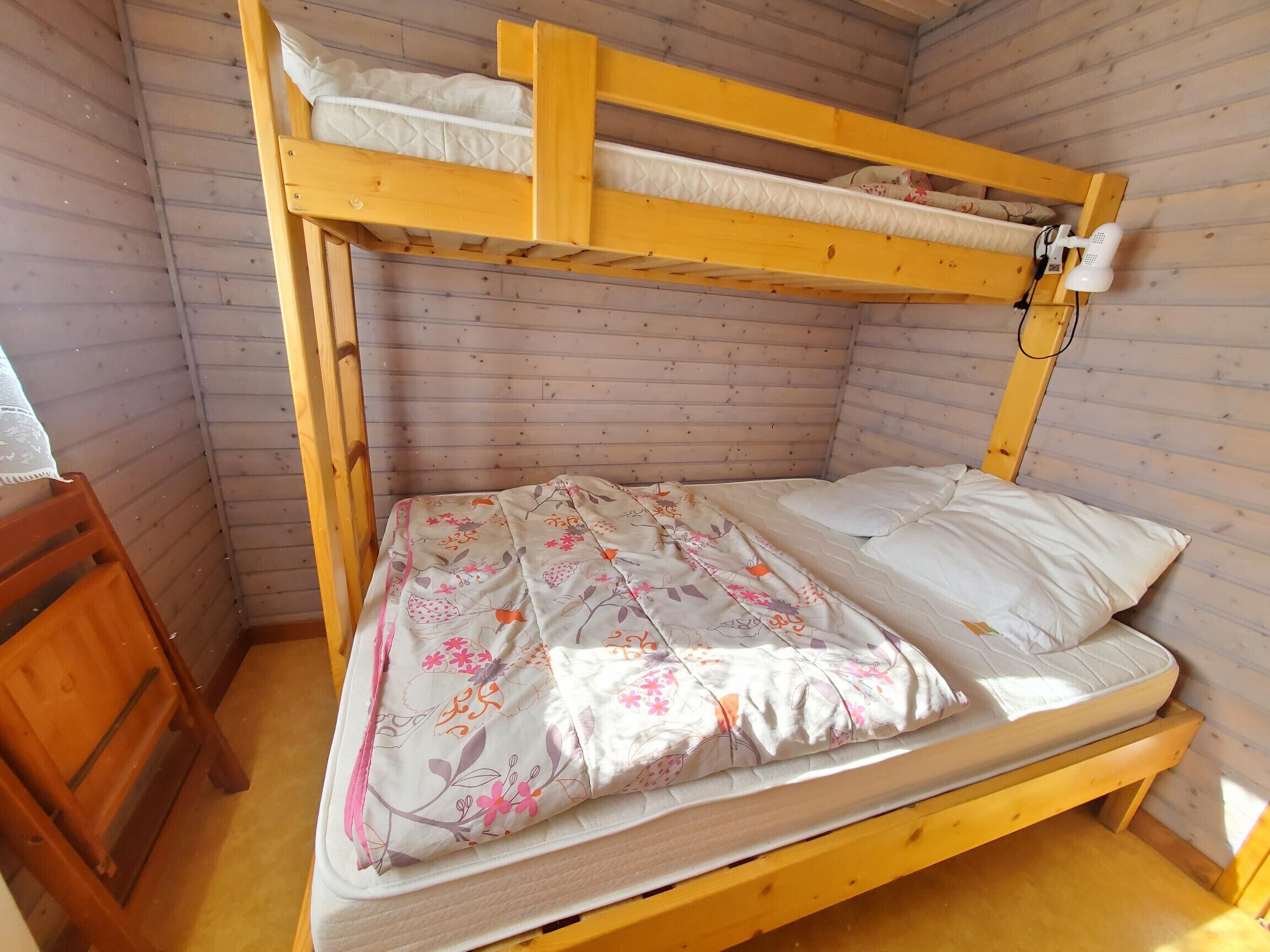 A bunk bed with a floral bedspread and pillows, wooden walls, and a small wooden chair.