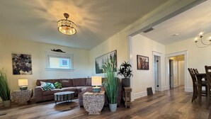 A spacious living room with a wooden floor, a brown leather sofa, a round coffee table, and a chandelier. There are two side tables with lamps and a large potted plant. - Charming Vintage Retreat in the Heart of Anaconda. (Anaconda)