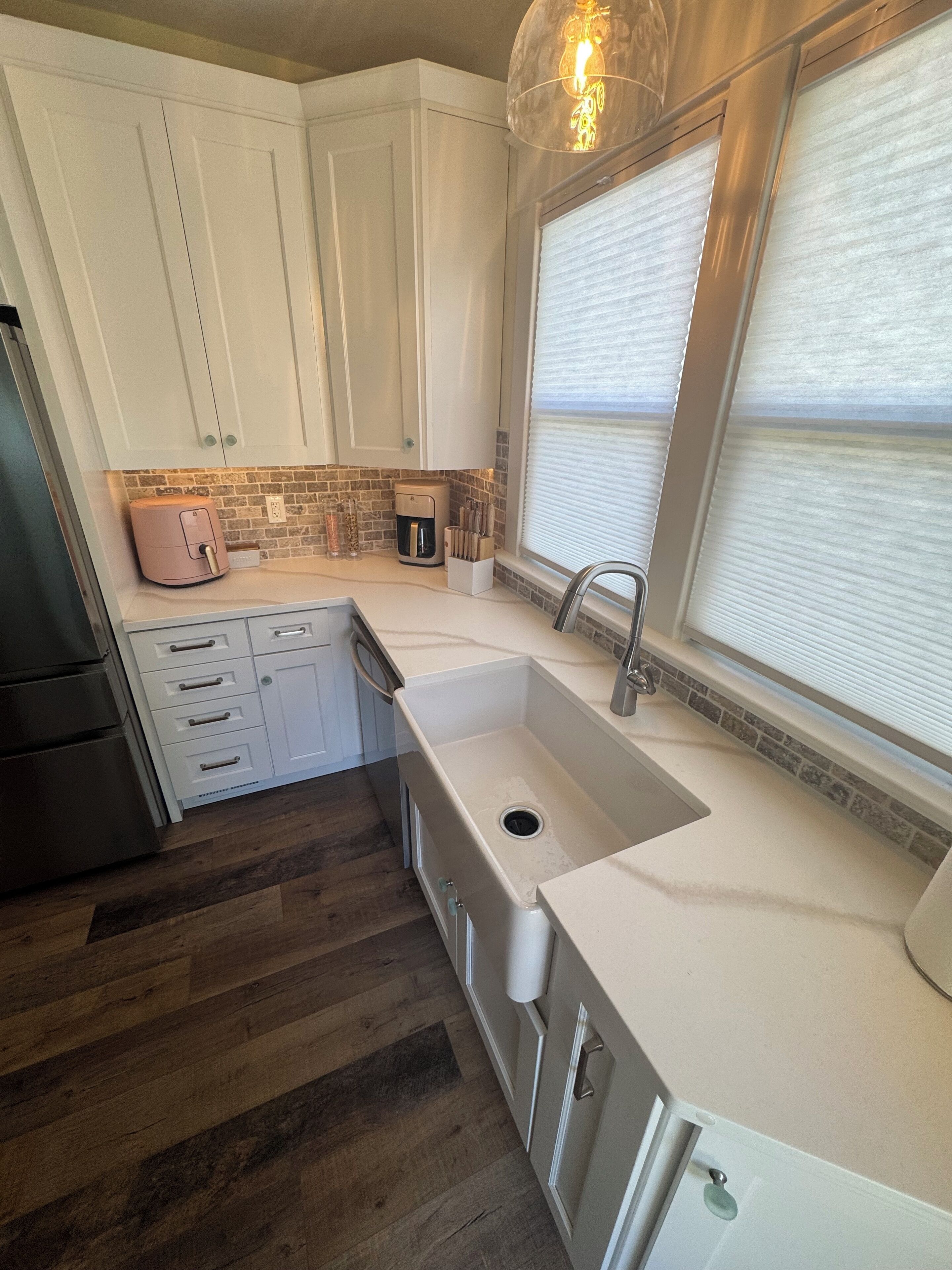 Una cocina con armarios blancos, un refrigerador de acero inoxidable, una cafetera rosa y una ventana con persianas.