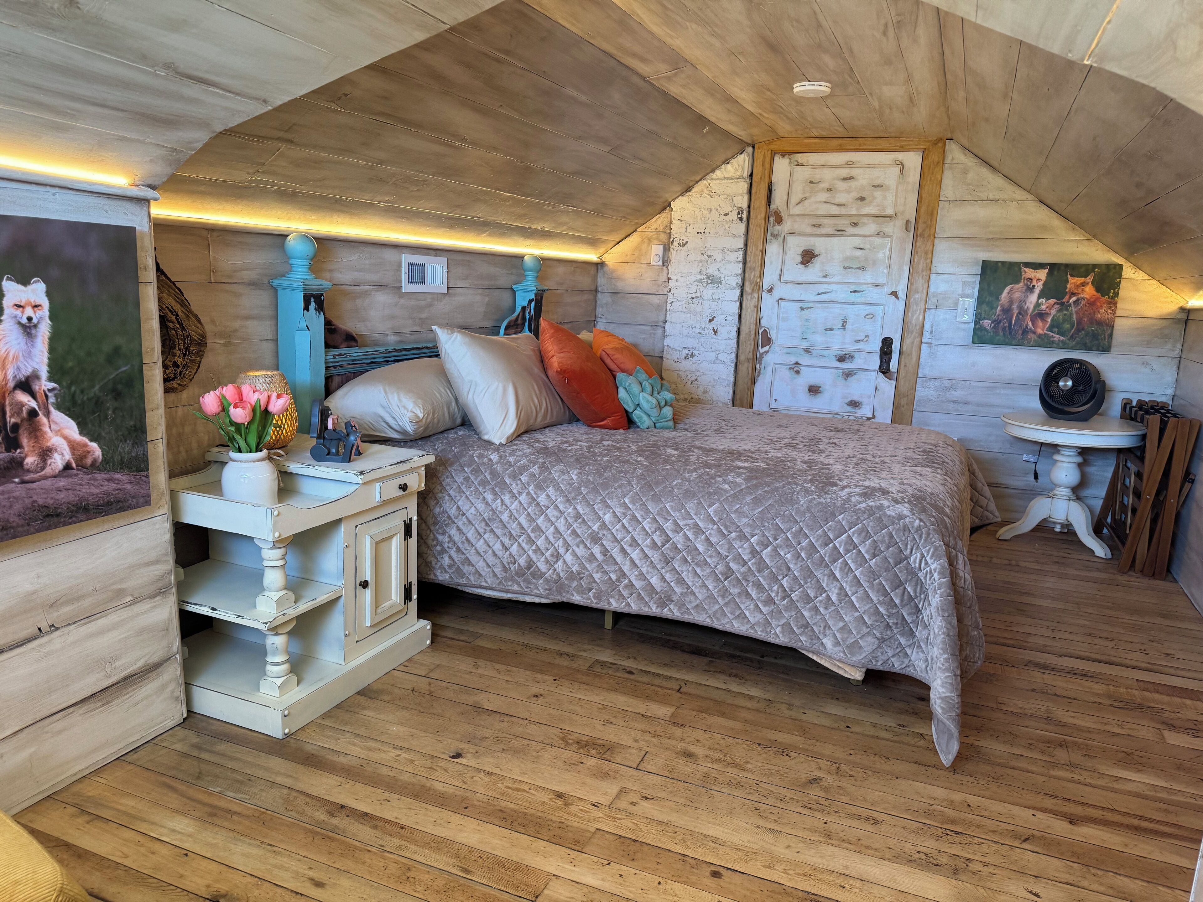 Une chambre confortable située sous les toits, avec un lit recouvert d’une courtepointe, un plancher en bois, une commode blanche et une petite table sur laquelle est posé un ventilateur.