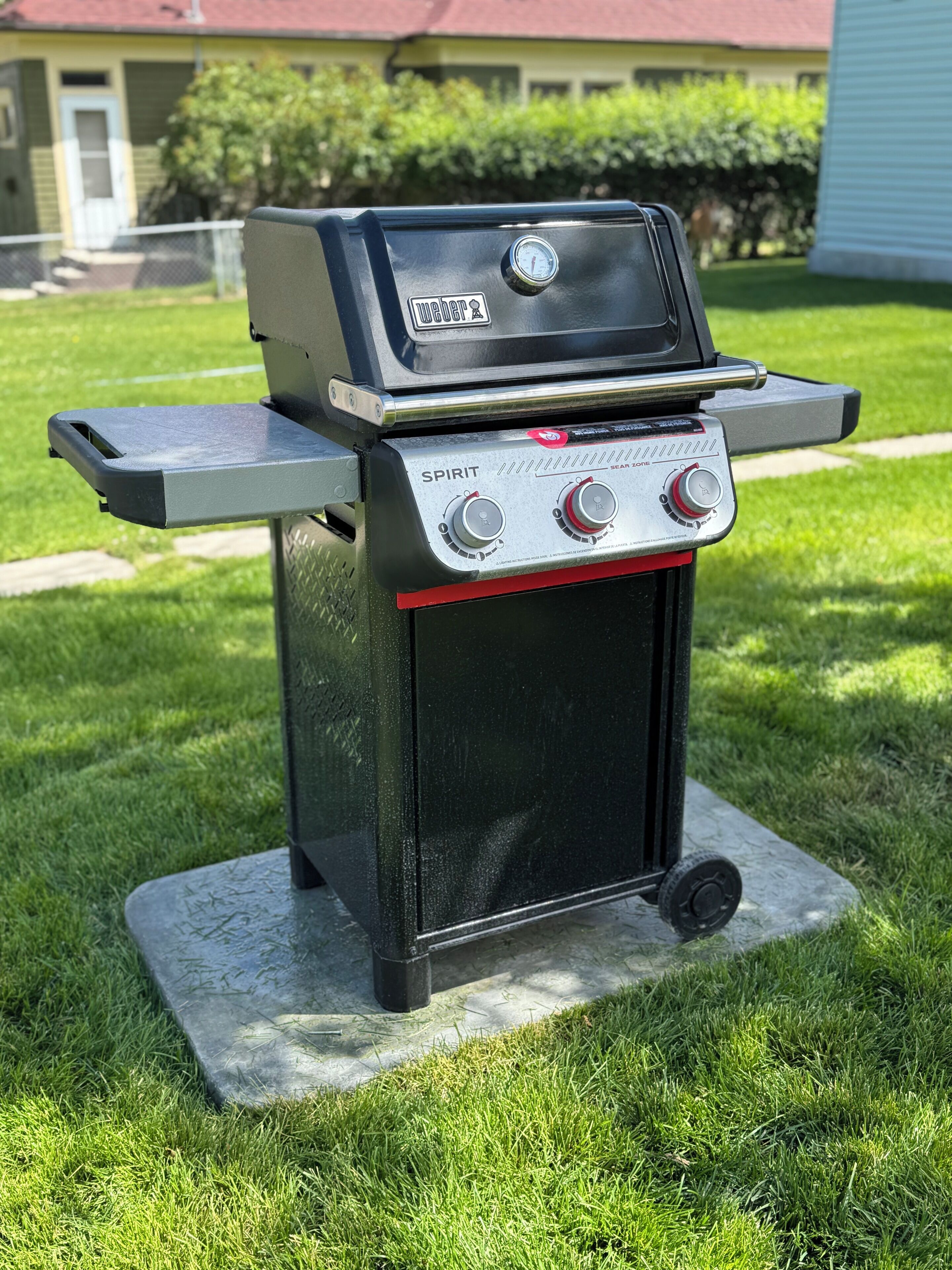 En svart utomhusgrill med en digital termometer och två justeringsknappar, placerad på en betongplatta i en gräsig trädgård.