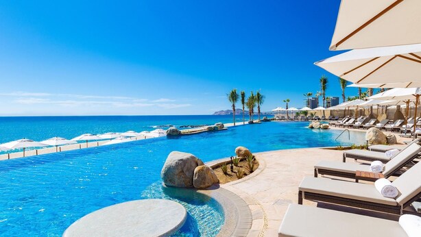 Zona de piscina en un resort con sillones de descanso y sombrillas, cielo azul despejado y vista al océano.
