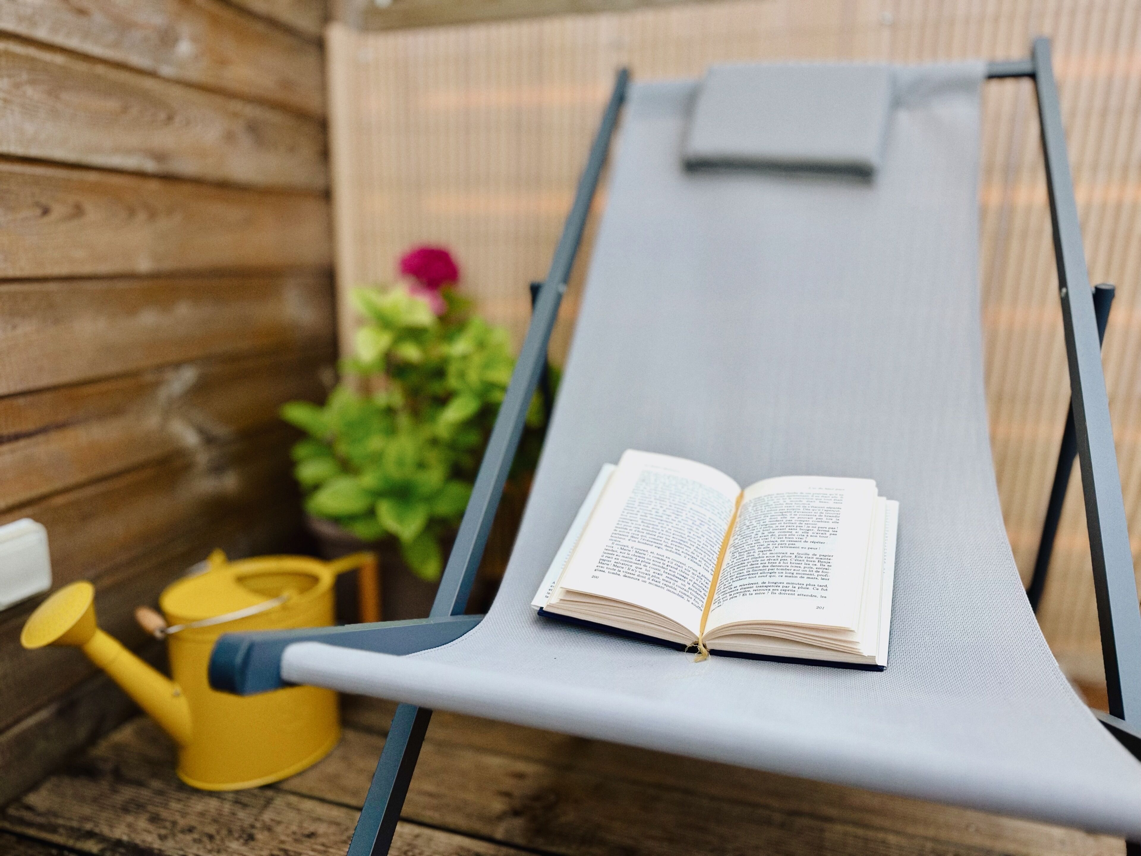 Une terrasse en bois avec un fauteuil de jardin gris, un livre ouvert et un arrosoir jaune.