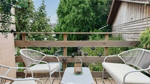 Un terrazzo in legno con sedie in vimini bianco e un tavolo, circondato da vegetazione e un recinto di legno.