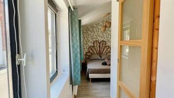 A narrow hallway with a bed in the background, wooden doors, and a window with a curtain.