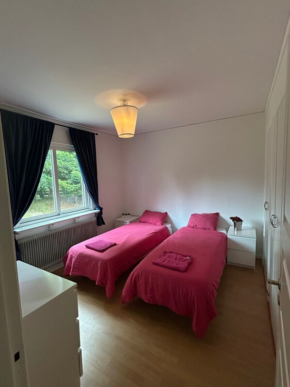 A bedroom with two beds, a window with curtains, a radiator, a bedside table with a lamp, and a white wardrobe.