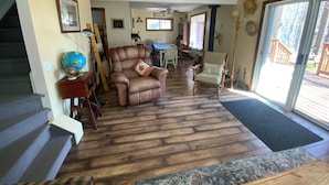 Amplia sala de estar con un sofá de cuero marrón, una silla de madera y una mesa de comedor con sillas. Se observa una escalera a la izquierda y un ventanal grande a la derecha.