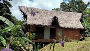 Une maison à toit de chaume avec une terrasse en bois, entourée d’une végétation luxuriante et de plantes en fleurs.
