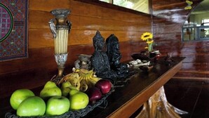 Une table en bois sur laquelle se trouve un panier de fruits contenant des pommes vertes, des bananes et des pommes rouges, ainsi qu’un vase décoratif et une petite statue.