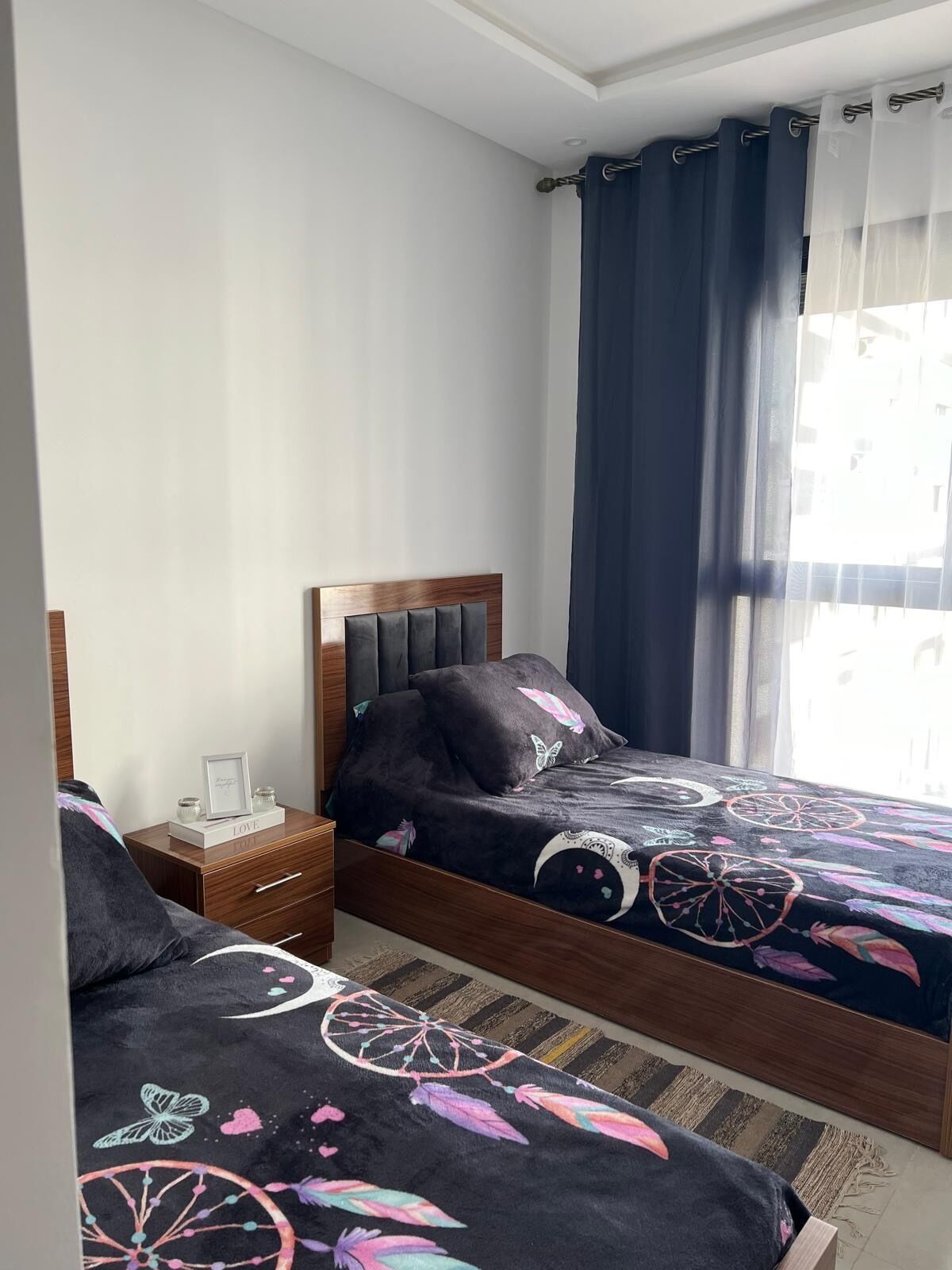 Une chambre avec deux lits, une tête de lit en bois, une table de chevet et une fenêtre avec des rideaux.