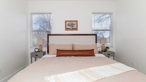 Ein Schlafzimmer mit einem großen Bett, zwei Nachttischen mit Lampen, einem gerahmten Bild an der Wand und Fenstern mit Jalousien.